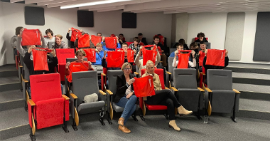 Zdjęcie przedstawia grupę uczniów wraz z dwoma nauczycielkami. Wspólnie pozują do zdjęcia z czerwonymi torbami materiałowymi firmy ROCKWOOL. Wszyscy znajdują się w małej sali konferencyjnej.