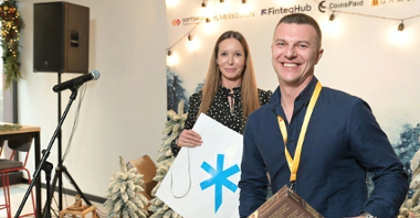 Zdjęcie przedstawia Ivana Montika, założyciela firmy SOFTSWISS. Mężczyzna jest uśmiechnięty, trzyma w ręku grę planszową "Łamacze szyfrów". Za nim stoi dyrektor Biura Obsługi Inwestorów Katja Lożina. W tle znajduje się świąteczna ścianka z logotypami firm.