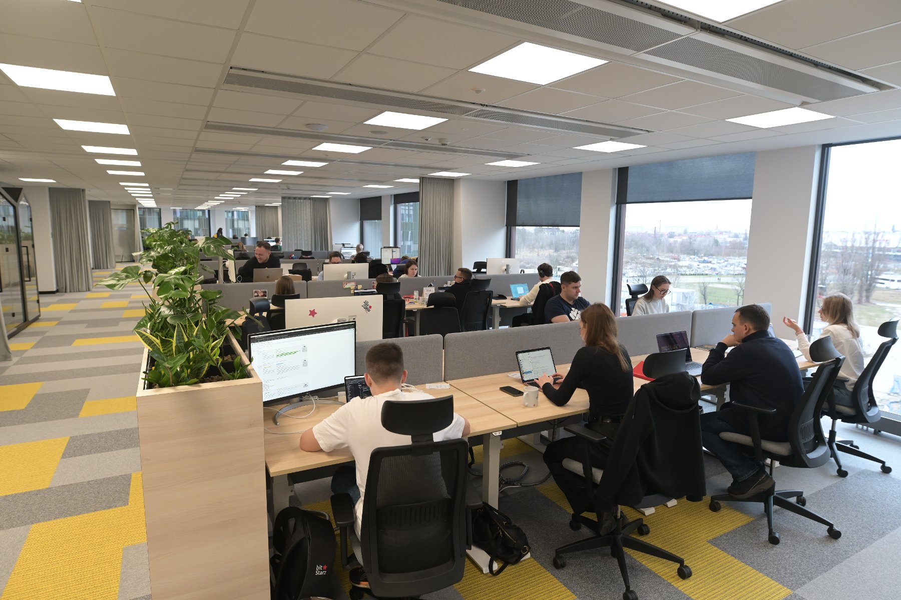 Zdjęcie przedstawia otwartą przestrzeń biurową. Wzdłuż ściany ciągną się biurka z komputerami, pracują przy nich osoby. - grafika artykułu