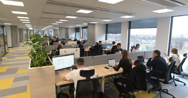 Zdjęcie przedstawia otwartą przestrzeń biurową. Wzdłuż ściany ciągną się biurka z komputerami, pracują przy nich osoby.