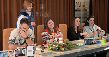 Zdjęcie przedstawia pracowników firmy SOFTSWISS w trakcie świątecznych warsztatów. Osoby siedzą przy stole, przygotowują stroiki świąteczne. Na stole stoją kleje i farbki, pędzelki, igliwie. Osoby śmieją się, są zadowolone.