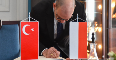 Na zdjeciu mężczyzna wpisujący się do księgi pamiątkowej, obok niej stoją dwie flagi: polska i turecka